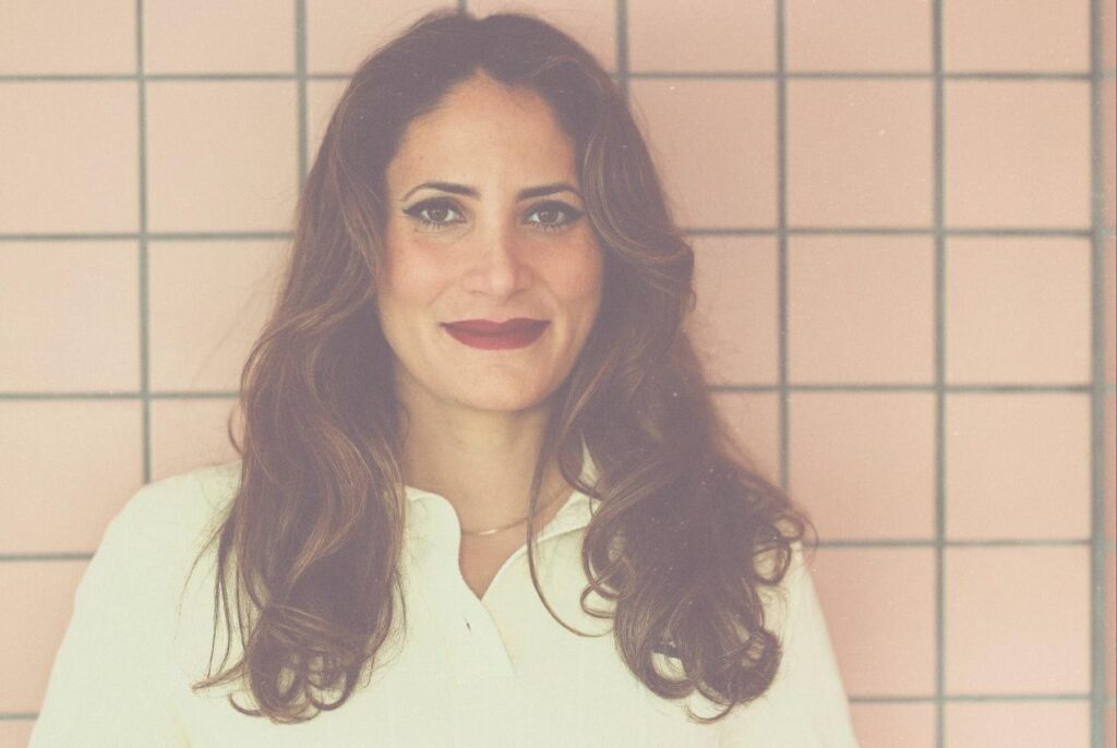 Eda Hamacher in einer weißen Bluse steht vor einer rosa Kachelwand. Sie hat schulterlange brauen HAare und lächelt in die Kamera